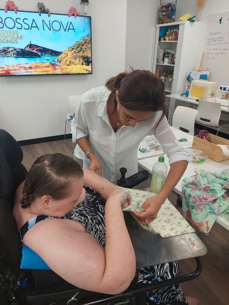 A woman is assisting a young person in a wheelchair with a craft activity. They are both focused on the task at hand, which involves fabric and a table surface. The woman is leaning in to help, while the young person attentively follows along. In the background, a screen displays the words 'Bossa Nova,' and the setting appears to be a bright, well-equipped room designed for creative or educational activities.