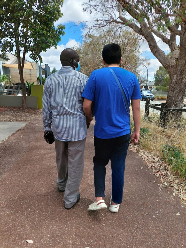 Two men walking side by side on a paved path, seen from behind. The man on the left is wearing a striped shirt and grey trousers, while the man on the right is wearing a blue t-shirt and dark pants. They appear to be walking together in a relaxed manner, possibly having a conversation. The setting is outdoors, with trees and a residential area visible in the background