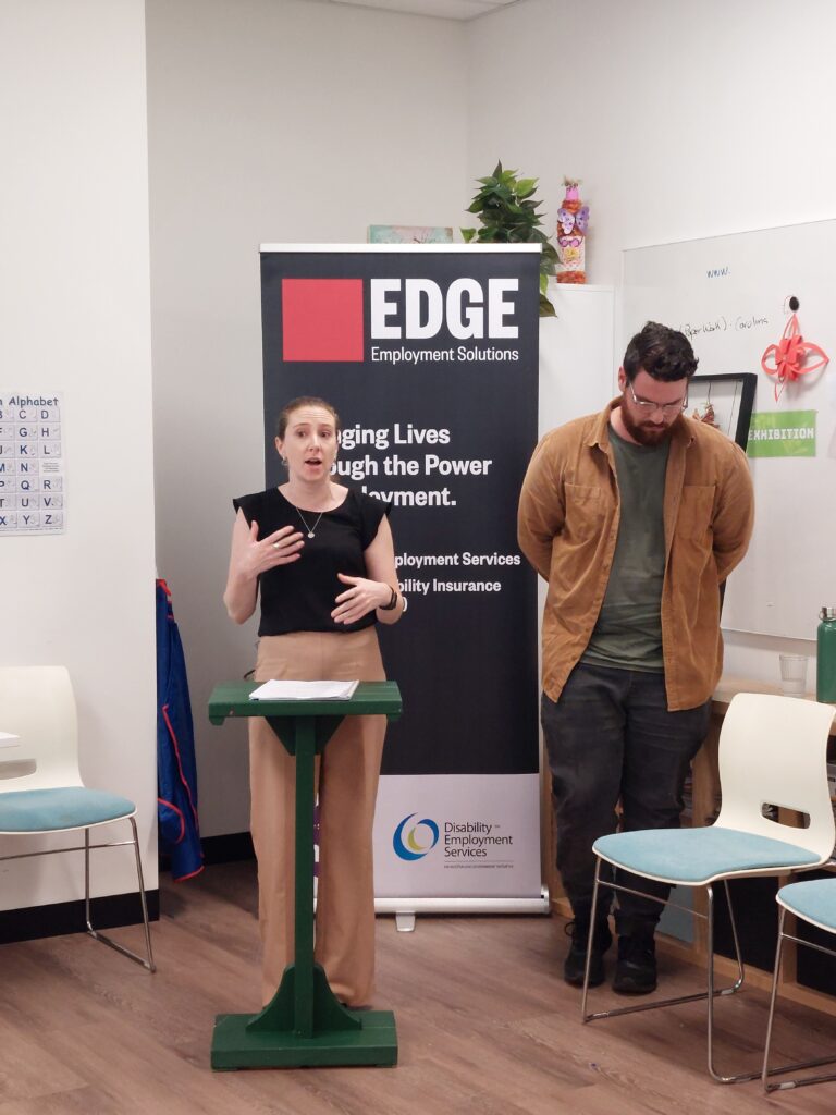 A woman standing at a small podium speaking to an audience, with a man standing beside her, looking down. Behind them is a banner with the logo 'EDGE Employment Solutions' and the tagline 'Changing Lives Through the Power of Employment.' The setting appears to be an indoor event or presentation space, with chairs and informational displays in the background