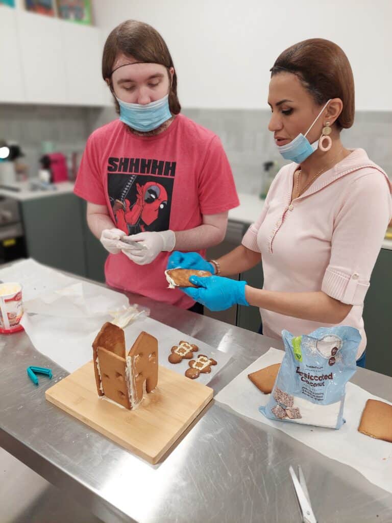 A young man and a woman wearing face masks and gloves are assembling a gingerbread house together in a kitchen. The young man is wearing a red t-shirt with a graphic design, and the woman is dressed in a light pink top. They are both focused on decorating the gingerbread house, with various decorating supplies on the stainless steel counter in front of them