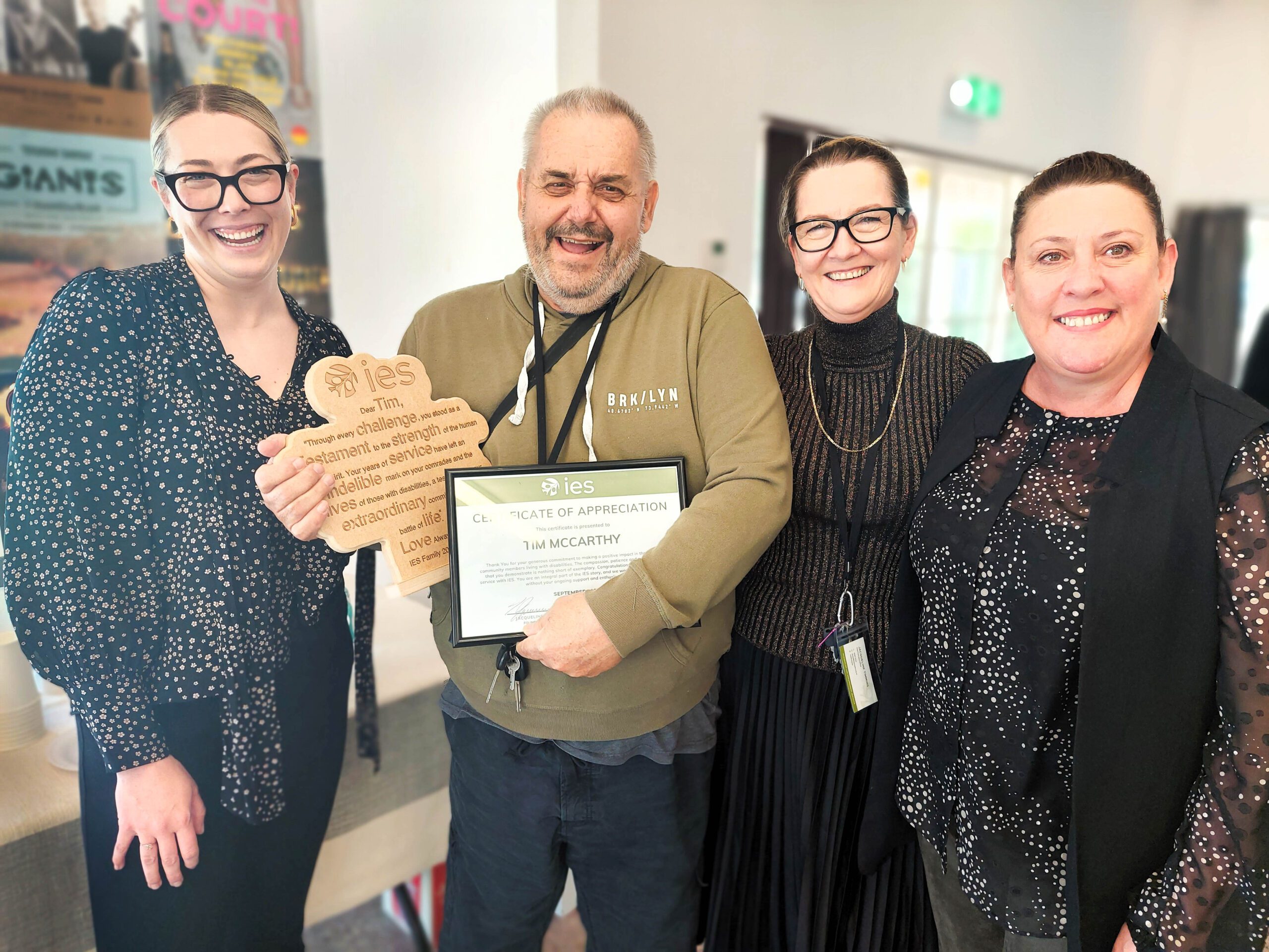 A group of four people are standing together indoors, smiling at the camera. The person in the center is holding a certificate of appreciation with the “IES” logo and their name, “Tim McCarthy.” They are also holding a wooden plaque shaped like a jigsaw puzzle piece, which has a heartfelt message of appreciation engraved on it. The two women on either side of Tim are dressed in black outfits, while the woman on the far left is wearing a black and white polka-dot blouse and glasses. The background includes some posters and a doorway.