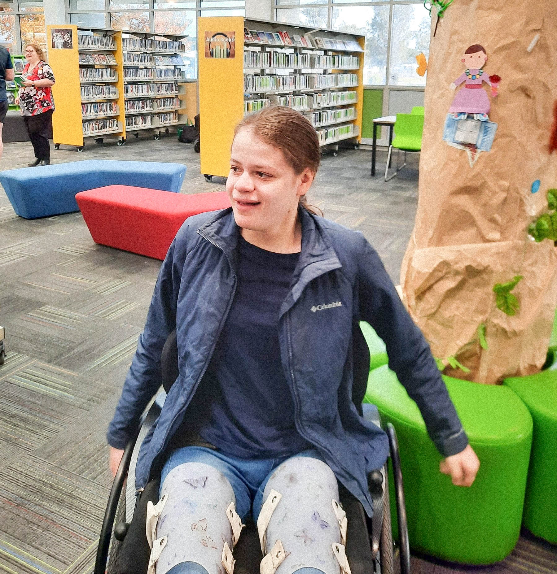 A young woman is sitting in a wheelchair inside a library. She is wearing a blue jacket, dark blue shirt, and light blue leggings with a butterfly pattern, along with leg braces. She has a cheerful expression on her face. In the background, there are bookshelves filled with books, colourful seating, and a large decorative tree made from paper. Another person is visible in the distance, browsing the shelves. The library environment appears bright and welcoming.