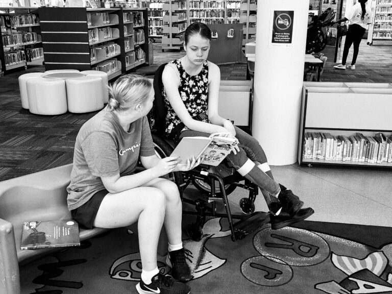 Two people are seated in a library, engaged in reading a book together. The person on the right is sitting in a wheelchair, wearing a floral-patterned top and jeans, while the person on the left is sitting on a small purple chair, wearing a blue T-shirt and shorts. The person on the left is holding the book open, and they are both focused on it. The library background includes bookshelves filled with books, colourful seating, and a vibrant rug with playful designs. The atmosphere is calm and educational.