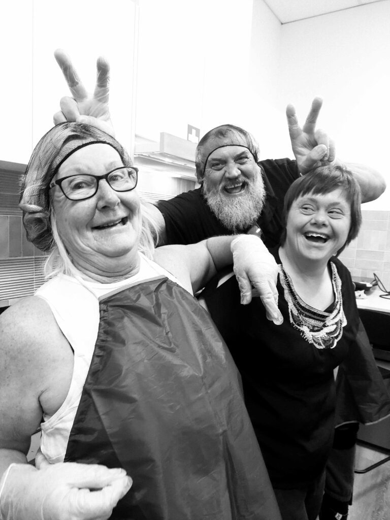 Three people are standing together in a kitchen, all smiling and enjoying themselves. The person on the left, an older woman wearing glasses, a blue apron, and a hairnet, has a playful expression as the man in the middle, who has a beard and is also wearing a hairnet, makes bunny ears behind her head. The person on the right, a young woman with short hair, is laughing and wearing a black top with a beaded necklace. All three are wearing disposable gloves, indicating they might be involved in a cooking activity. The atmosphere is fun and lighthearted.