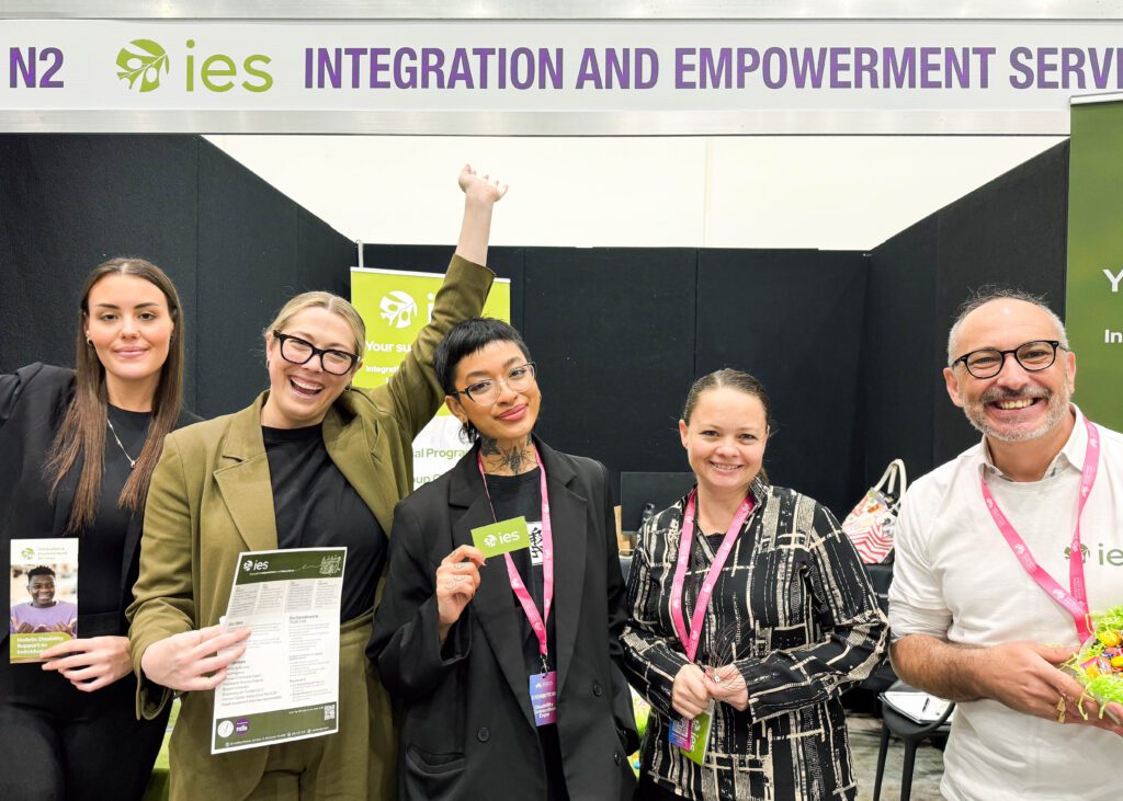 A group of five people stand together at an expo booth under a banner that reads “IES Integration and Empowerment Services.” They are all smiling and holding various promotional materials. The woman second from the left is raising her hand in celebration. The individuals are dressed in professional attire, and the booth behind them features IES branding, including a logo and informational banners. The mood is positive and energetic, reflecting the team’s enthusiasm at the event.