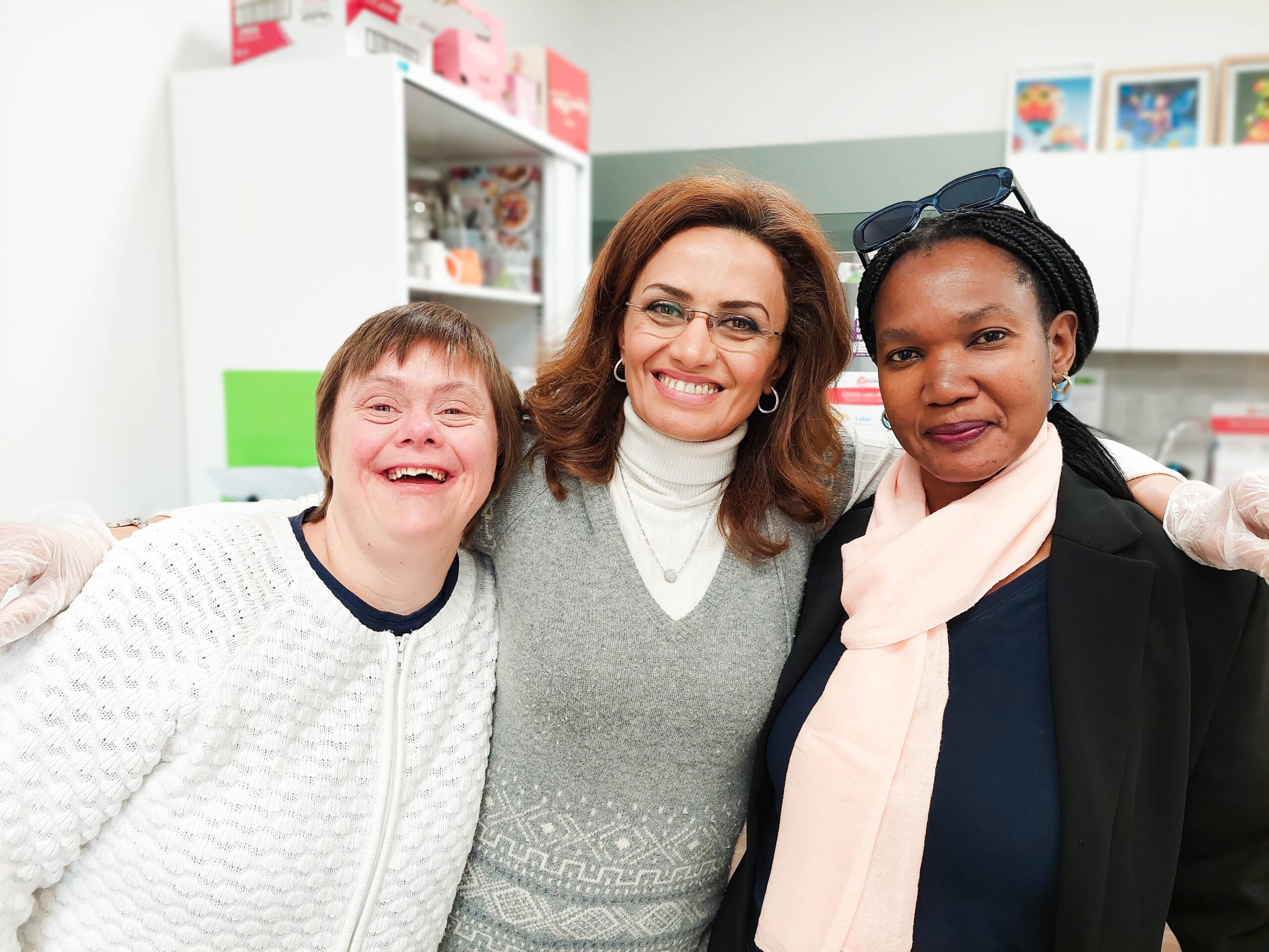 Three women are standing close together indoors, smiling warmly at the camera. The woman on the left has short hair and is wearing a white textured sweater. The woman in the center has medium-length brown hair, is wearing glasses, and is dressed in a grey sweater over a white turtleneck. The woman on the right has braided hair, is wearing a black blazer, a navy top, and a light pink scarf. The background includes white shelves and cabinets, with various items and decorations visible.