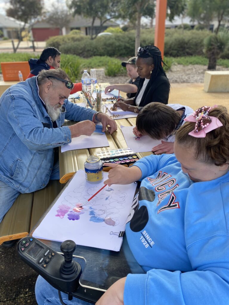 A group of people seated at an outdoor table, engaged in a painting activity. The individuals are focused on their artwork, using brushes and watercolours on paper. One person in the foreground is using a wheelchair and is painting with a brush. The setting appears to be a park or garden area with trees and greenery in the background, creating a relaxed and creative atmosphere.