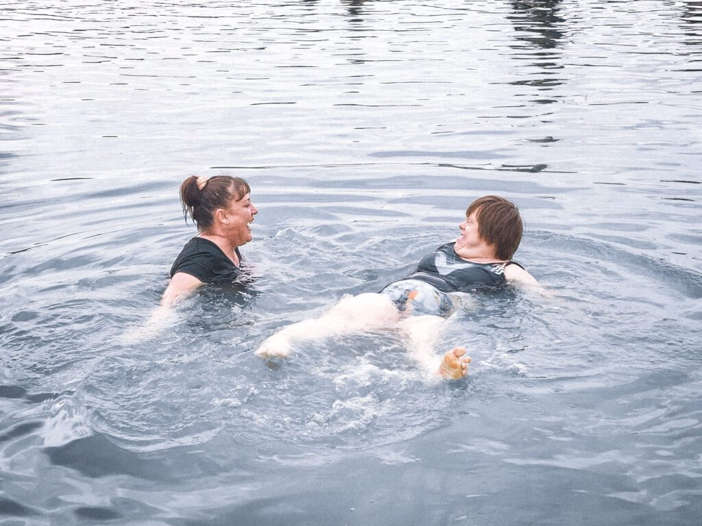 Two women are in the water, enjoying a swim together. The woman on the left, with her hair tied in a ponytail, is smiling and supporting the other woman, who is floating on her back. The woman floating is wearing a colourful swimsuit, and both seem to be having a pleasant and relaxed time. The water is calm and clear, creating a peaceful and refreshing atmosphere. The scene captures a moment of enjoyment and care between the two women.