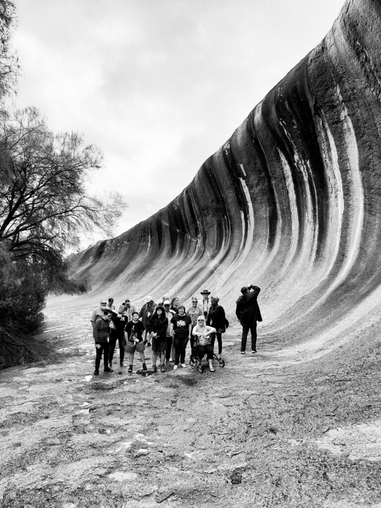 A group of people are standing together at the base of Wave Rock, a famous natural rock formation in Australia known for its wave-like shape. The group includes both standing individuals and one person in a wheelchair. The sky is overcast, and the massive, curved rock formation towers above them, showcasing streaks of different colours along its surface. The surrounding area is rocky with sparse vegetation. The group appears to be on a guided tour or outing, enjoying the scenic and iconic location despite the cloudy weather.